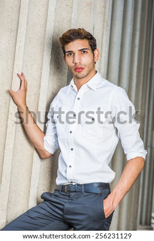 Portrait of Young Professional. Wearing a white shirt, sleeves rolling over, black pants, a young college student with a little beard is standing by columns on campus, confidently looking forward.