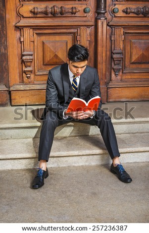 Man casual reading. Dressing in three pieces black suit, patterned necktie, vest, leather shoes, a young handsome college student is sitting on stairs in the door way, looking down, reading red book.