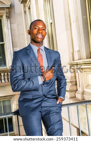 Successful businessman. Dressing in blue suit, tie, putting his hand on chest, a young black guy sitting on railing in vintage style office building, smiling, looking at you. Instagram filtered look.