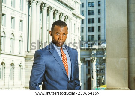 Businessman thinking hard. Dressing in blue suit, patterned tie, a young black guy standing in the front of vintage style office building, frowned, unhappily looking at you. Instagram filtered look.