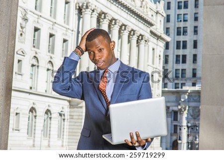 Businessman thinking hard. Dressing in blue suit, patterned tie, a young black guy standing in the front of vintage style office building, frowned, hand touching his head, working on laptop computer.