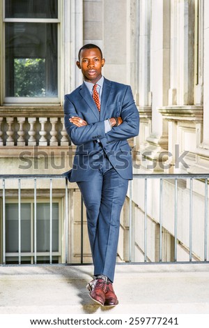 Man Urban Fashion. Dressing in blue suit, tie, leather shoes, a young black businessman sitting on railing in vintage style office building, crossing arms, legs, relaxing. Instagram filtered look.