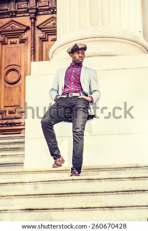 Urban fashion. Wearing newsboy cap, dressing in blazer, patterned under shirt, pants, leather shoes, a young black guy standing stairs outside vintage style office building. Urban retro filtered look.