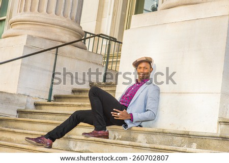 Man Urban Fashion. Wearing newsboy cap, dressing in light gray blazer, patterned pink, black under shirt, black pants, brown leather shoes, a young black guy sitting on stairs, looking at you.