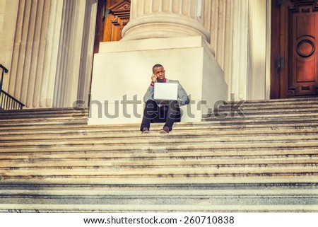 Way to Success. A black business man sitting on stairs of vintage style office building door way, working on laptop computer and talking on his mobile phone in the same time. Instagram filtered look.