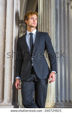 Portrait of successful businessman. Dressing in black suit, necktie, a young sexy businessman with curly hair is standing by column outside vintage style office building, confidently looking forward.