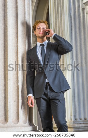 Businessman calling anywhere. Dressing in black suit, necktie, a young sexy guy with curly hair standing by column outside vintage style office building, listening, talking to his mobile phone.