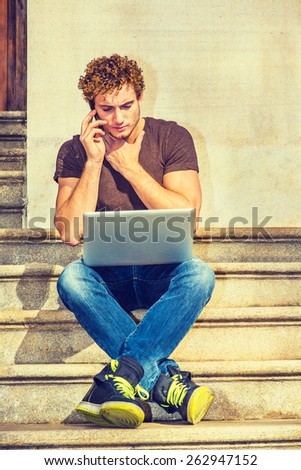 A young guy sitting on stairs outside office, crossing legs, working on laptop computer, talking on phone, a hand covering the throat - a difficult, frustrated, long conversation. Instagram effect.