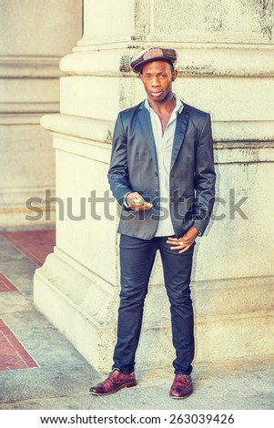 Wearing fashionable Newsboy cap, dressing in black blazer, pants, brown leather shoes, a young black guy standing by column on street, holding a mobile phone, waiting your message. Instagram effect.