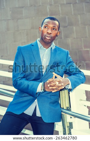 Portrait of Business Man. Wearing blue blazer, black pants, short haircut, ear stud, bracelets, finger ring, a young black guy standing against metal fences by wall, looking forward to meeting you.