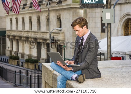 Young businessman working on street. Young blonde, handsome student, wearing blazer, necktie, jeans, crossing legs, looking dow, working on laptop computer. Wall Street sign, flags on background.