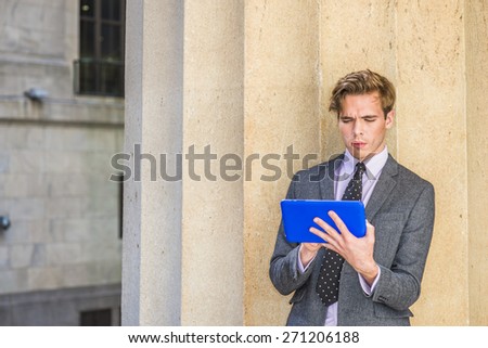 Young blonde, handsome student, wearing blazer, necktie, standing by column outside office, hands holding a tablet computer, typing, reading, thinking. Concept of new technology in our daily life.