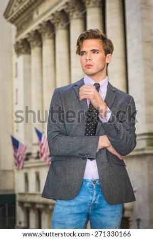 Portrait of Businessman. Young blonde, handsome graduate student, wearing blazer, necktie, jeans, standing by vintage style office building with American flags, looking forward. Urban casual fashion.