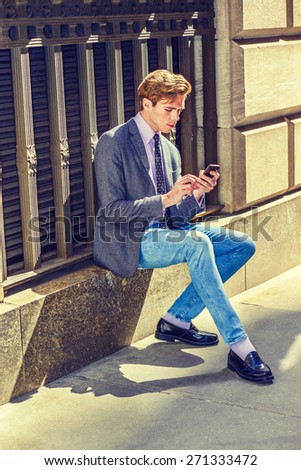 Texting under sunshine. Young blonde, handsome student, wearing blazer, necktie, jeans, jeans, sitting on vintage style window on street, hands typing messages on his mobile phone. City Daily life.