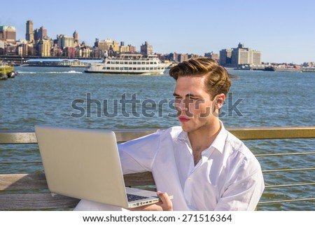 Study Outside. Young blonde, handsome student, wearing white shirt, sitting by river, reading, working on laptop computer. a small boat, big city on far background. Traveler.