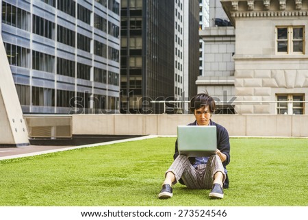 Japanese student studying in New York. Wearing patterned shirt, black sweater, casual pants, leather shoes, a young guy sitting on green lawn in business district, reading, working on laptop computer.