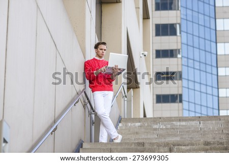 Man working remotely. Young blonde professional, wearing red sweater, white pants, reading, working on laptop computer, standing by wall outside office building. Concept of Technology in daily life.