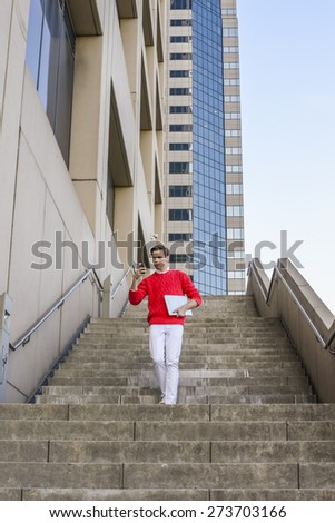 Young blonde professional, wearing red sweater, white pants, holding laptop computer, reading messages on his mobile phone, walking down stairs outside office building. Technology in daily life.