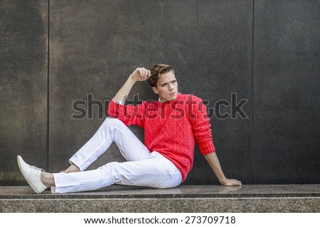 Man Urban Casual Fashion. Young blonde professional, wearing red sweater, white pant, casual fashion shoes, bending a leg and a arm, sitting on a marble bench, against the wall, relaxing, thinking.