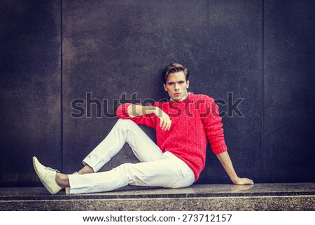Man Urban Fashion. Young blonde professional, wearing red sweater, white pants, fashion shoes, bending leg, arm resting on knee, sitting on a marble bench, against the wall. Instagram filtered effect.