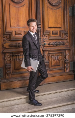Portrait of School Boy. Dressing formally in black suit, necktie, white shirt, carrying laptop computer, a young college student walking down stairs form vintage style office door way on campus, smile