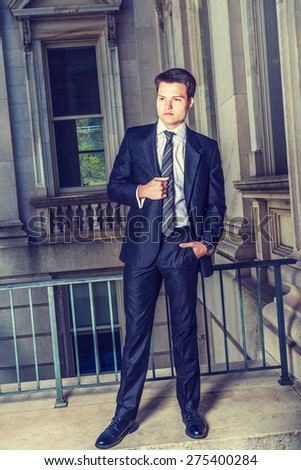 Dressing in black suit, necktie, white shirt, leather shoes, a young college student standing by railing in vintage style office building on campus, looking forward. Instagram filtered effect.