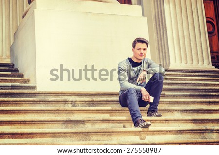 Student on Campus. Wearing gray V neck cardigan sweater, blue pants, brown sneaker, wristwatch, holding mobile phone, a young guy sitting on stairs outside office building, smiling, looking at you.