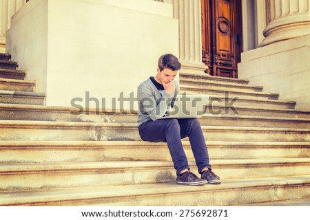 Wearing gray V neck cardigan sweater, blue pants, brown sneaker, a young college student sitting on stairs outside office building, hand touching chin, looking down, working on laptop computer.