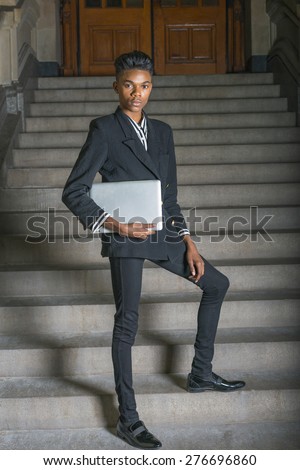 School boy.  A young dark skin, 18 years old student, wearing black fashionable blazer, pants, leather shoes, holding laptop computer, standing on stairs outside office, confidently looking at you.