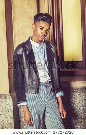 Portrait of School Boy. A young,18 years old student, wearing black fashionable jacket, blue dyed white shirt, striped pants, standing by window, inclining head, interestedly looking at you.