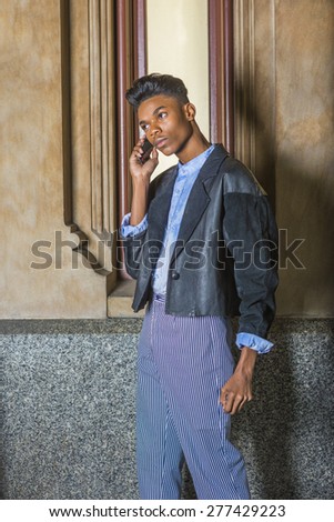 Technology in Daily Life. A young, 18 years old student, wearing black fashionable jacket, blue dyed white shirt, striped pants, standing by window on campus, making phone call on his mobile phone.