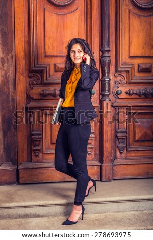 Dressing in black blazer, orange shirt, pants, heels, a young East Indian American business woman walking down vintage style office door way, carrying laptop computer, making call on her mobile phone.