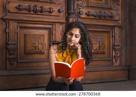 Way to Success. Power of Reading. A young beautiful East Indian American college student sitting by vintage style library door way, tilting head, hand touching chin, looking down, reading red book.