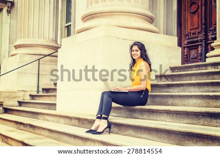 Wearing sleeveless orange shirt, striped pants, high heels, a young East Indian American college student sitting on stairs outside office building on campus, smiling, working on laptop computer.
