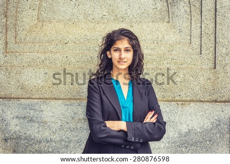 Modern East Indian American Student. Dressing in black blazer, blue under shirt, crossing arms,  a young girl with long curly hair standing by wall, smiling, looking at you. Instagram filtered effect.