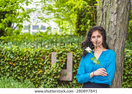 A flower girl. Wearing long sleeve blue shirt, holding a white rose, a young East Indian American college student standing by tree in spring day, waiting for you. A love, passion, romantic girlfriend.