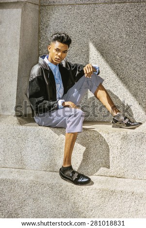 Urban teenage fashion. A 18 years old black student, wearing fashionable jacket, striped pants, blue dyed white shirt, leather shoes, sitting by rocky wall under sun shine, thinking. Instagram effect.