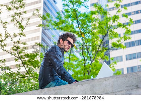 Wearing leather jacket, sunglasses, a young handsome guy with beard, reading, working on laptop computer on the top of wall on business district in spring day. Technology in our daily life.
