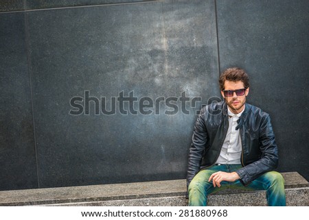 Dressing in black leather jacket unbuttoned, white under shirt,  blue jeans, wearing sunglasses, a young guy with beard sitting on marble bench against wall on street, sad, thinking. Copy Space.
