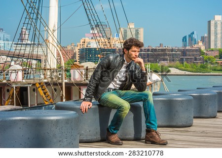 Man Spring Casual Urban Fashion. Wearing black leather jacket, blue jeans, brown leather boot shoes, a young guy with beard, sitting on bench at harbor, relaxing, thinking. A boat on background.