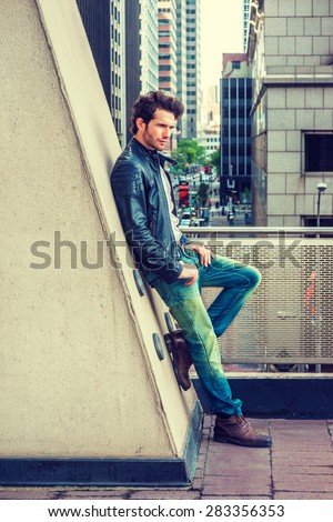 Man Urban Casual Fashion. Wearing black leather jacket, blue jeans, brown boot shoes, a young guy with beard, leaning against wall on balcony, facing street, thinking. City Boy. Instagram effect.