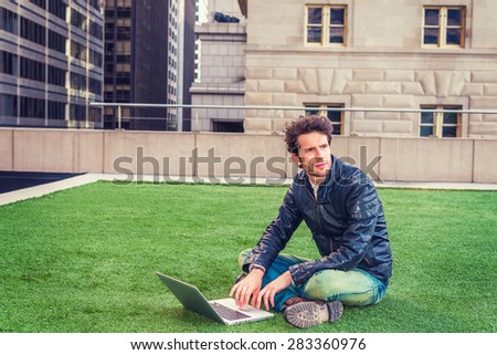 European graduate student studying in New York. Wearing black leather jacket, blue jeans, boot shoes, young guy with beard, crossing legs, sitting on green lawn, working on laptop computer, thinking.
