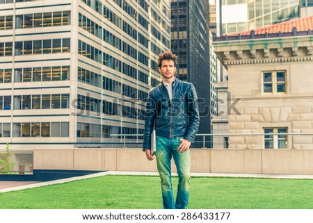 European graduate student in New York. Wearing black leather jacket, blue jeans, carrying laptop computer, a young guy with beard, standing on lawn in business district, confidently looking forward.