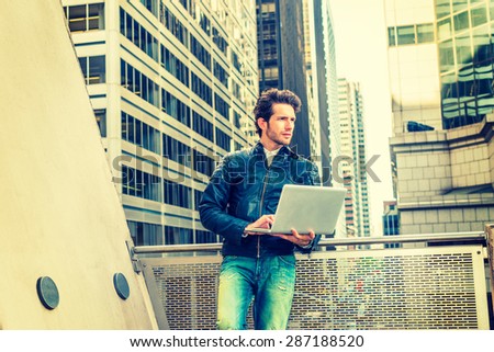 European businessman working in New York. Wearing black leather jacket, jeans, young guy with beard, standing by railing on balcony in business district, working on laptop computer. Instagram effect.