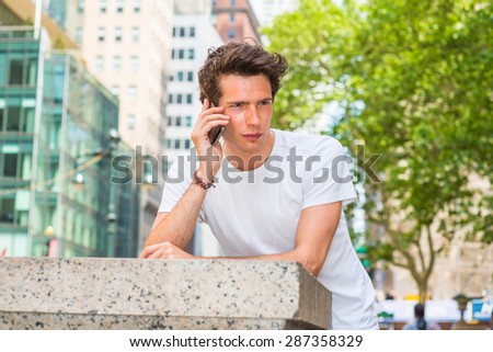European college student traveling in New York. Wearing white T shirt, a young guy looking down, thinking, calling on his mobile phone outside on street in summer. Technology in our daily life.