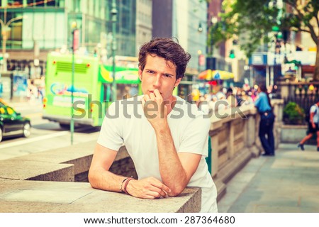 Portrait of Innocent Young College Student. Wearing white T shirt, hand touching his lips, a young European guy standing by stone fence on street in New York, watching, thinking. Instagram effect.