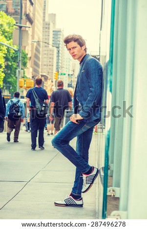 European College Student in New York. Wearing blue blazer, jeans, sneakers, holding laptop computer, a young guy standing against glass wall on street. People walking on background. Instagram effect.