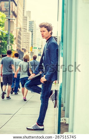 Wearing blazer, jeans, sneakers, holding laptop computer, a young European student standing against glass wall on street in New York, feeling lonely. Young couple holding hands, walking on background.