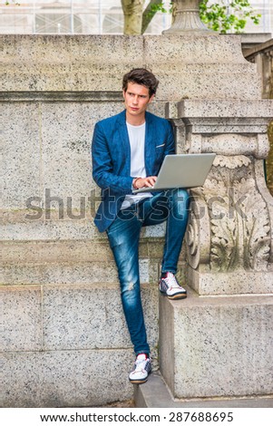 European college student studying in New York. Wearing blue blazer, jeans, sneakers, a young guy staying at quiet corner on campus, working on laptop computer, reading, thinking. Working Anywhere.