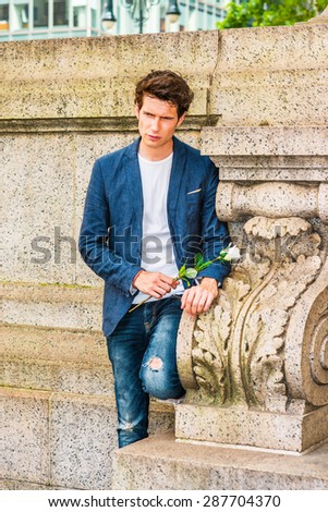 European college student seeking love in New York. Wearing blue blazer, jeans, sneakers, a young guy standing at corner against wall on campus, holding white rose, missing for you. Instagram effect.